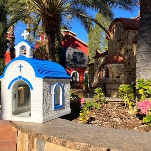 Puede incluir: Una peque&ntilde;a r&eacute;plica de capilla blanca y azul con un frente de vidrio y un techo azul. La capilla est&aacute; sentada en un muro de piedra frente a un edificio de piedra m&aacute;s grande con un techo rojo.