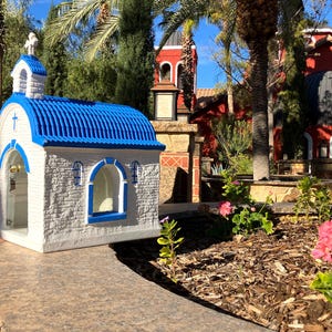 Puede incluir: Una peque&ntilde;a r&eacute;plica de capilla blanca y azul con un techo azul y una cruz en la parte delantera. La capilla est&aacute; rodeada de plantas verdes y un edificio rojo en el fondo.