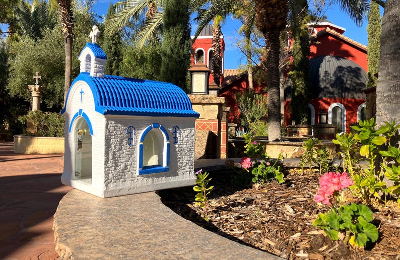 Puede incluir: Una peque&ntilde;a r&eacute;plica de capilla blanca y azul con un techo azul y una cruz en la parte delantera. La capilla est&aacute; rodeada de plantas verdes y un edificio rojo en el fondo.
