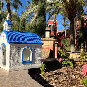 Puede incluir: Una peque&ntilde;a capilla en miniatura blanca y azul con un techo azul y una cruz en la parte superior. La capilla est&aacute; frente a un edificio rojo m&aacute;s grande con una c&uacute;pula.
