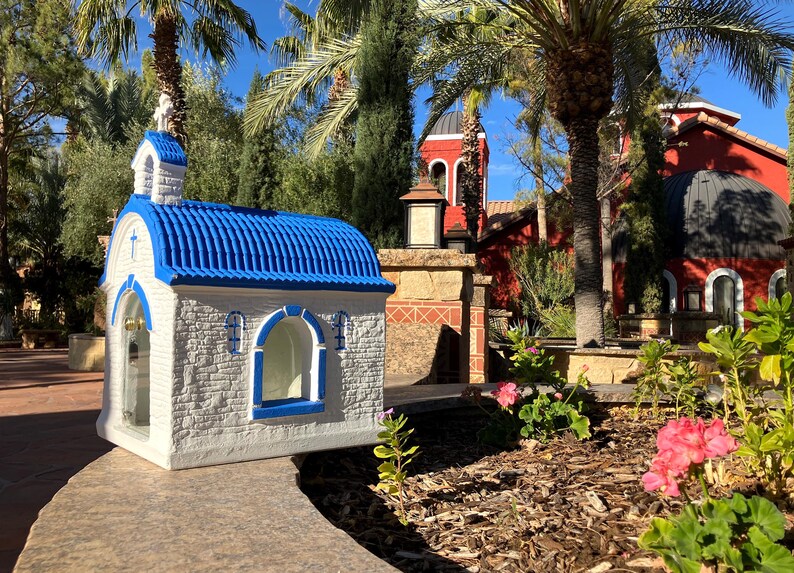 Puede incluir: Una peque&ntilde;a capilla en miniatura blanca y azul con un techo azul y una cruz en la parte superior. La capilla est&aacute; frente a un edificio rojo m&aacute;s grande con una c&uacute;pula.