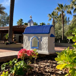 Puede incluir: Una peque&ntilde;a r&eacute;plica de capilla blanca y azul con un techo azul y una cruz blanca en la parte superior. La capilla est&aacute; rodeada de plantas verdes y palmeras.