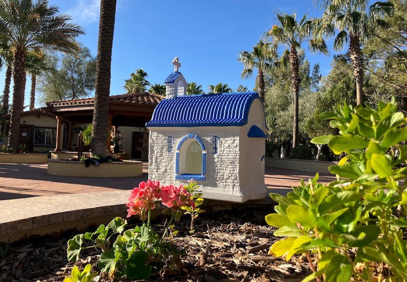 Puede incluir: Una peque&ntilde;a r&eacute;plica de capilla blanca y azul con un techo azul y una cruz blanca en la parte superior. La capilla est&aacute; rodeada de plantas verdes y palmeras.