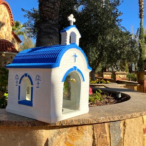 Puede incluir: Una peque&ntilde;a r&eacute;plica de capilla blanca y azul con un techo azul y una cruz en la parte delantera. La capilla tiene una ventana de vidrio y un campanario. La capilla est&aacute; sentada en una pared de piedra.