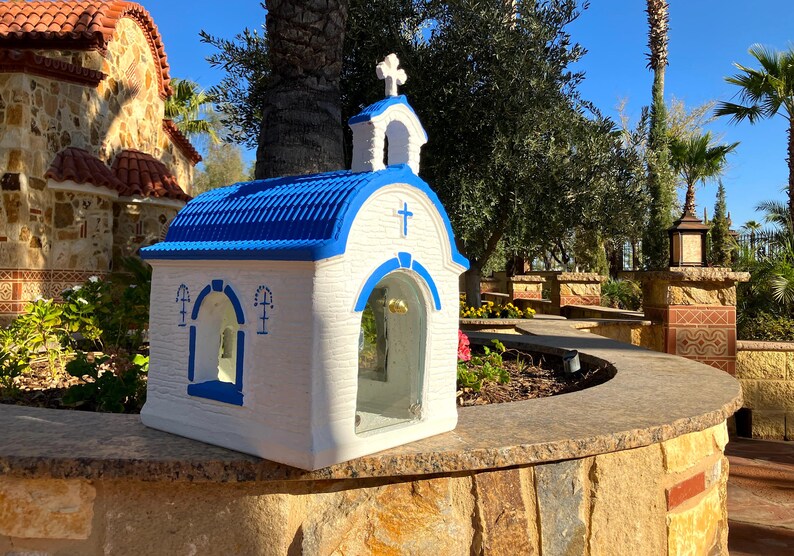 Puede incluir: Una peque&ntilde;a r&eacute;plica de capilla blanca y azul con un techo azul y una cruz en la parte delantera. La capilla tiene una ventana de vidrio y un campanario. La capilla est&aacute; sentada en una pared de piedra.