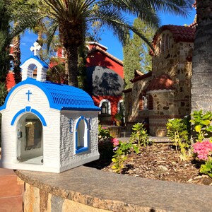 Puede incluir: Una peque&ntilde;a r&eacute;plica de capilla blanca y azul con un frente de vidrio y un techo azul. La capilla est&aacute; sentada en un muro de piedra frente a un edificio de piedra m&aacute;s grande con un techo rojo.