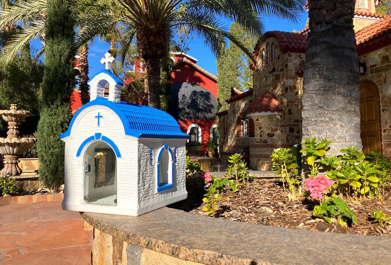 Puede incluir: Una peque&ntilde;a r&eacute;plica de capilla blanca y azul con un frente de vidrio y un techo azul. La capilla est&aacute; sentada en un muro de piedra frente a un edificio de piedra m&aacute;s grande con un techo rojo.