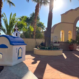 Puede incluir: Una peque&ntilde;a r&eacute;plica de capilla blanca y azul con un techo azul y una cruz en la parte superior. La capilla est&aacute; sentada sobre una superficie de piedra frente a un gran arco con una puerta de hierro forjado.