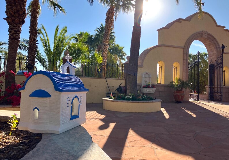 Puede incluir: Una peque&ntilde;a r&eacute;plica de capilla blanca y azul con un techo azul y una cruz en la parte superior. La capilla est&aacute; sentada sobre una superficie de piedra frente a un gran arco con una puerta de hierro forjado.