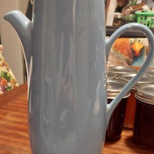 May include: A tall, light blue ceramic coffee pot with a white lid. The pot has a curved handle and a spout for pouring. The pot is sitting on a wooden surface.