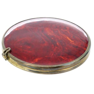 May include: A round, brass, hinged compact with a red marbled design under a clear glass top.