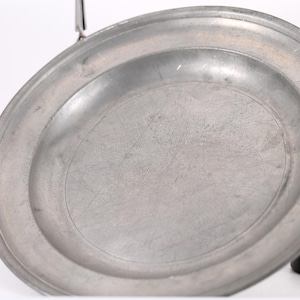 May include: A circular, metallic gray plate with a smooth, slightly reflective surface and a raised rim. The plate features visible surface scratches and a matte finish, held in place by a metal clamp attached to a black circular base.