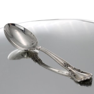 May include: A silver teaspoon with an ornate handle. The spoon is on a reflective surface.