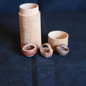 May include: A collection of small, handcrafted wooden items. The image features a tall, cylindrical container, a small cup, and three miniature basket-shaped objects. The items are made of light brown wood and sit on a dark surface.
