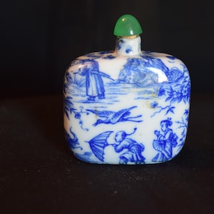 May include: A small, rectangular white ceramic snuff bottle featuring intricate blue painted illustrations of figures, a flying crane, and foliage. The bottle is topped with a smooth, rounded green stopper that sits atop a narrow neck.