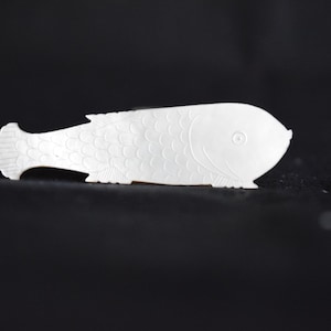 Könnte beinhalten: Ein weißer, geschnitzter, fischförmiger Perlmutt-Anhänger.