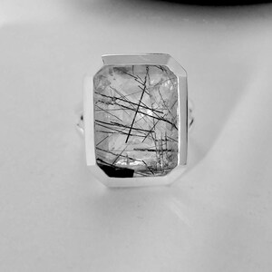 Puede incluir: Un anillo de plata con una piedra de cuarzo transparente con inclusiones negras. La piedra est&aacute; engastada en un bisel cuadrado.