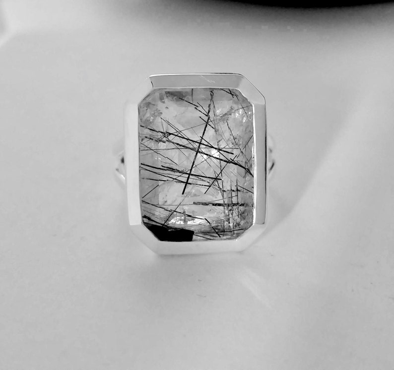 Puede incluir: Un anillo de plata con una piedra de cuarzo transparente con inclusiones negras. La piedra est&aacute; engastada en un bisel cuadrado.
