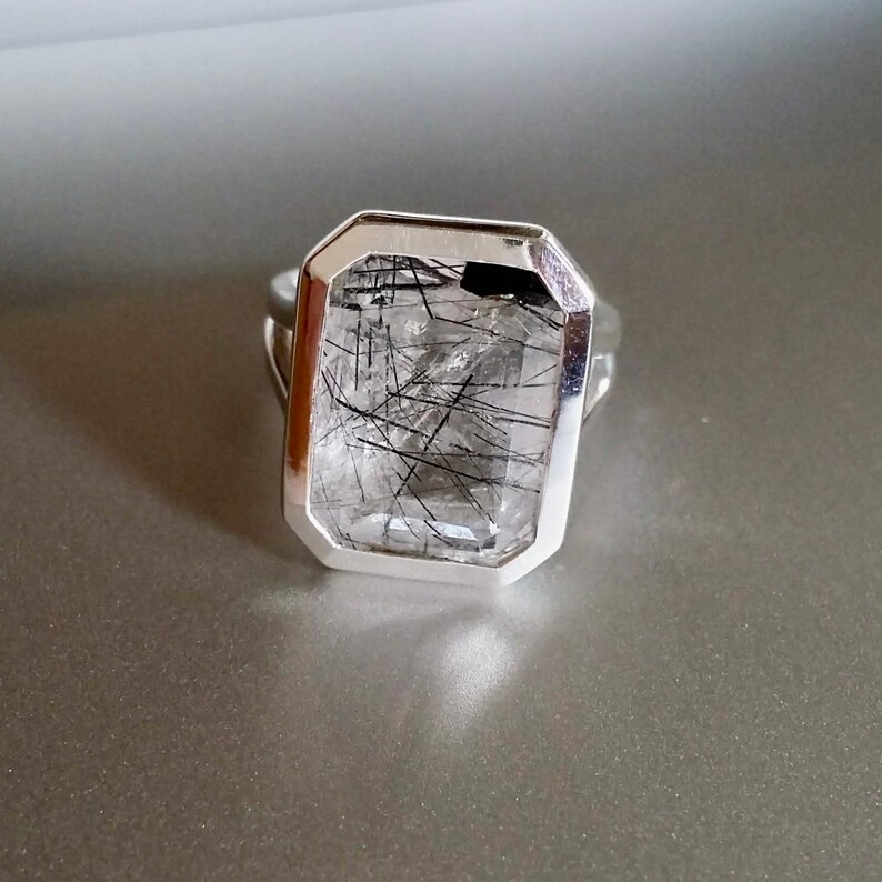 Puede incluir: Un anillo de plata con una piedra preciosa de cuarzo transparente con inclusiones negras. La piedra preciosa est&aacute; engastada en un bisel rectangular.