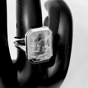 Puede incluir: Un anillo de plata con una piedra preciosa rectangular transparente con inclusiones negras. La piedra preciosa est&aacute; engastada en un engaste de bisel.