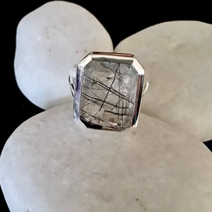 Puede incluir: Un anillo de plata con una piedra preciosa de cuarzo transparente que presenta inclusiones negras. La piedra preciosa est&aacute; tallada en forma rectangular con bordes biselados.