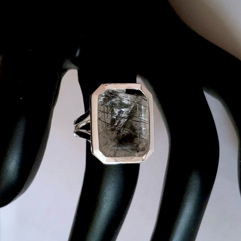 Puede incluir: Un anillo de plata con una piedra grande de cuarzo rectangular transparente con inclusiones negras. La piedra est&aacute; engastada en un engaste de bisel.