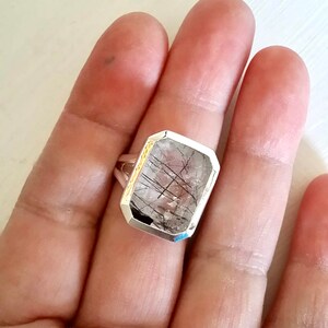 Puede incluir: Un anillo de plata con una piedra de cuarzo transparente con inclusiones negras. La piedra est&aacute; tallada en forma rectangular con bordes biselados.