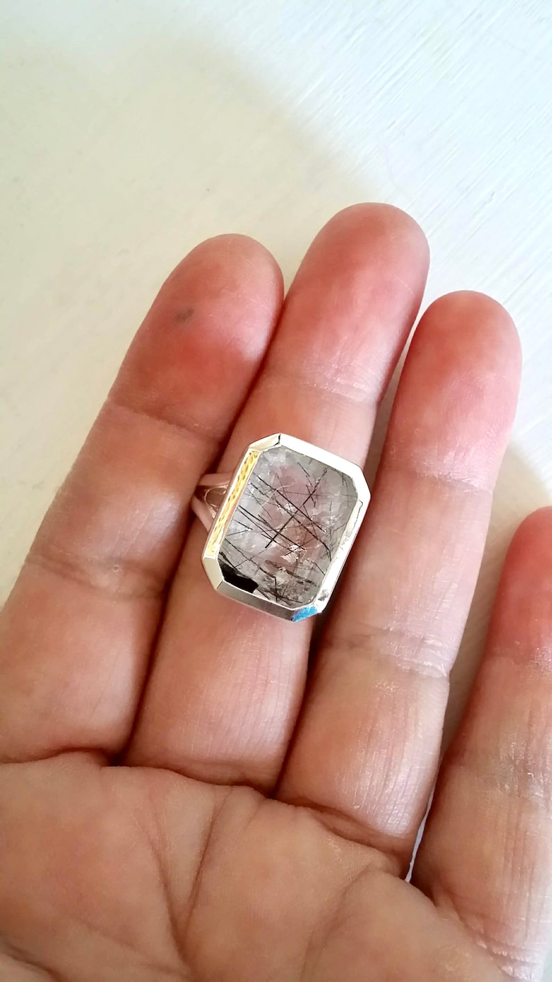 Puede incluir: Un anillo de plata con una piedra de cuarzo transparente con inclusiones negras. La piedra est&aacute; tallada en forma rectangular con bordes biselados.