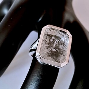 Puede incluir: Un anillo de plata con una piedra preciosa rectangular transparente que tiene inclusiones negras. La piedra preciosa est&aacute; engastada en un engaste de bisel.