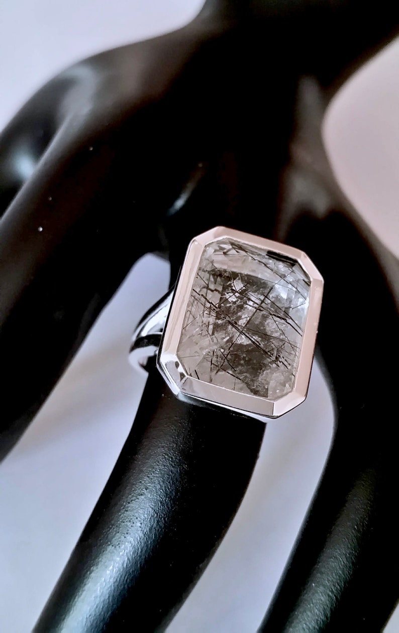 Puede incluir: Un anillo de plata con una piedra preciosa rectangular transparente que tiene inclusiones negras. La piedra preciosa est&aacute; engastada en un engaste de bisel.