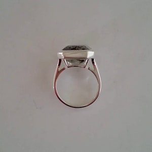Puede incluir: Un anillo de plata con una piedra preciosa transparente facetada engastada en un engaste cuadrado. La piedra preciosa tiene inclusiones negras.