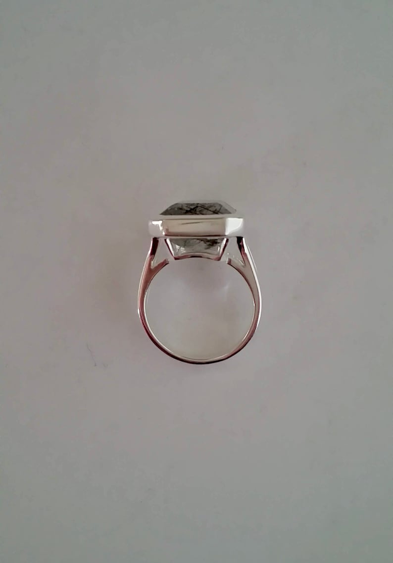 Puede incluir: Un anillo de plata con una piedra preciosa transparente facetada engastada en un engaste cuadrado. La piedra preciosa tiene inclusiones negras.