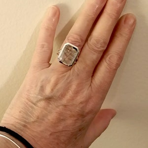 Puede incluir: Un anillo de plata con una piedra rectangular transparente que presenta inclusiones marrones. La piedra est&aacute; engastada en un engaste de bisel.