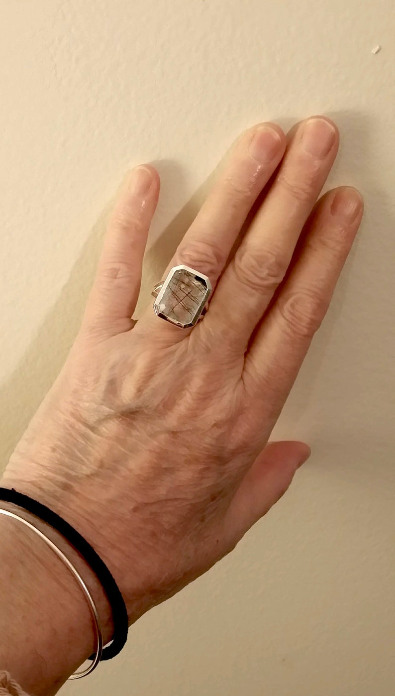 Puede incluir: Un anillo de plata con una piedra rectangular transparente que presenta inclusiones marrones. La piedra est&aacute; engastada en un engaste de bisel.