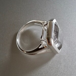 Puede incluir: Un anillo de plata con una gran piedra preciosa cuadrada transparente con inclusiones negras. La piedra preciosa est&aacute; engastada en un engaste de bisel.