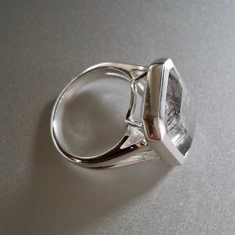 Puede incluir: Un anillo de plata con una gran piedra preciosa cuadrada transparente con inclusiones negras. La piedra preciosa est&aacute; engastada en un engaste de bisel.