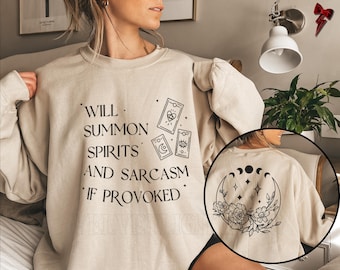 May include: A beige sweatshirt with the text "Will summon spirits and sarcasm if provoked" and a tarot card design. The back of the sweatshirt has a moon and flower design.