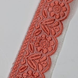 May include: Close-up of a coral-colored embroidered ribbon with a floral design. The ribbon features detailed stitching of flowers and leaves along the edge, with a white sheer fabric backing. The ribbon is suitable for crafts and sewing projects.