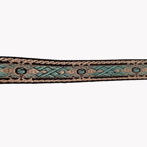 May include: A decorative woven trim featuring a repeating geometric pattern in shades of light turquoise and cream, bordered by thin black lines. The fabric ribbon displays a symmetrical design with diamond shapes and small circular accents throughout.