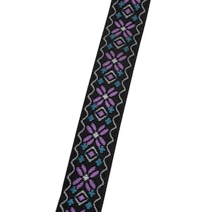 May include: Close-up of a black woven ribbon with a repeating floral pattern in shades of purple and teal. The ribbon has a wavy white border on each side, creating a decorative trim.