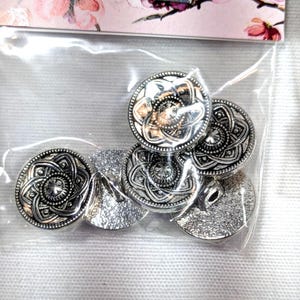 May include: A set of round, silver-toned metal buttons featuring an intricate, embossed floral pattern with a central star-like design. The buttons are packaged in a clear plastic bag, positioned below a card with a pink floral illustration.