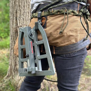 Puede incluir: Un escalón de soporte de árbol de metal gris está unido a un arnés de caza marrón y verde. El arnés se lleva alrededor de la cintura y se asegura con una cuerda. La persona lleva vaqueros grises oscuros.