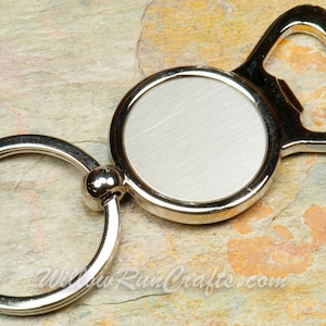 May include: Silver bottle opener keychain with a round, blank, metal tag and a key ring.