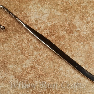 May include: A silver bookmark with a curved end and a hook at the top. The bookmark is on a brown textured surface.