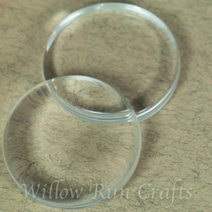May include: Two clear, circular glass pieces, possibly coasters or decorative elements, are displayed against a textured, neutral-toned background. The pieces are transparent, allowing the background to be visible through them. The text "Willow Run Crafts" is visible.