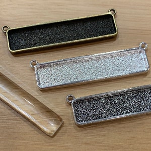 May include: Three rectangular metal blanks for jewelry making, one silver, one gold, and one black, and a clear glass rod.