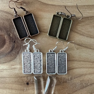 May include: Four pairs of silver, bronze, and copper rectangular earring blanks with clear glass inserts. The earring blanks have a textured surface.