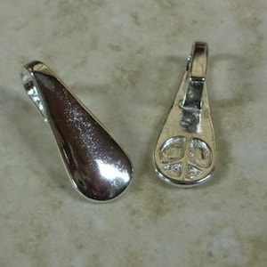 May include: Two silver-colored teardrop-shaped pendants. One pendant is smooth and reflective, while the other features a peace sign on the back. Both have a small loop at the top for attaching to a chain or cord. The pendants are displayed on a neutral-toned surface.