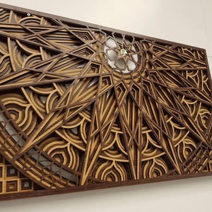 May include: Geometric wooden wall art with a layered design. The artwork features a complex pattern of shapes and lines in shades of brown and tan, creating a three-dimensional effect. The piece is framed in a dark brown wood.