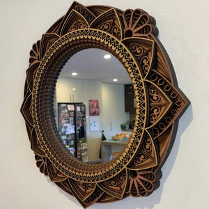 Könnte beinhalten: Ein runder, mehrschichtiger Holzspiegel mit einem Mandala-Design. Der Spiegel hat einen dunkelbraunen Rahmen mit aufwendigen Ausschnittmustern in Braun- und Beigetönen. Der Spiegel hängt an einer weißen Wand.